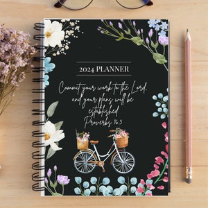 Op de afbeelding: Een 2024-planner met een zwarte kaft met bloemen aquarelontwerpen en een fietsillustratie. De tekst luidt: "Vertrouw uw werken aan de Heer toe, en uw plannen zullen slagen. Spreuken 16:3". Een potlood en een bril liggen op het houten oppervlak.