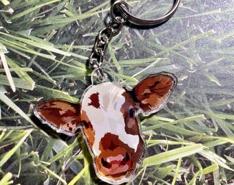 May include: A brown and white cow keychain with a silver key ring. The cow's face is visible and the keychain is on a green grassy background.