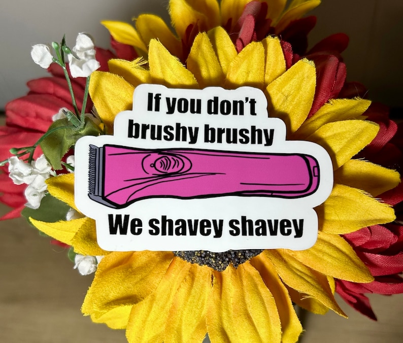 K&ouml;nnte beinhalten: Ein Aufkleber mit dem Text "If you don't brushy brushy We shavey shavey" und einer rosa Haarschneider-Illustration. Der Aufkleber befindet sich auf einem Strau&szlig; Sonnenblumen mit roten und gelben Bl&uuml;tenbl&auml;ttern.