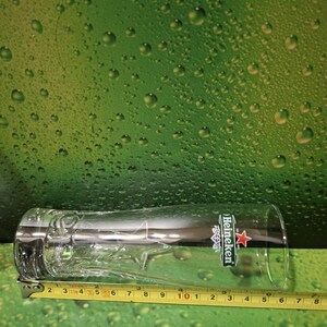 May include: A clear, tall beer glass with the Heineken logo. The glass has a decorative star design near the base and is approximately 20 cm long. The glass is on a green background.