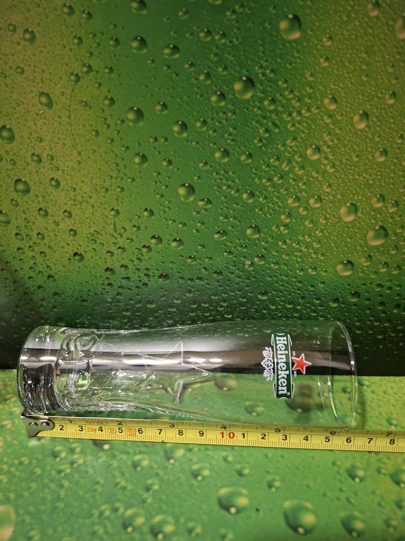 May include: A clear, tall beer glass with the Heineken logo. The glass has a decorative star design near the base and is approximately 20 cm long. The glass is on a green background.