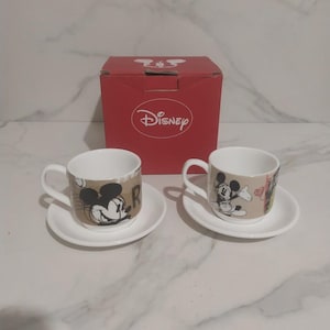 Op de afbeelding: Een set van twee witte keramische espressokopjes met schotels, met Mickey Mouse-ontwerpen. De kopjes hebben een bruine band met Mickey Mouse-afbeeldingen en de letter "R". Een rode Disney-doos staat op de achtergrond.