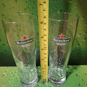 May include: Two clear Heineken beer glasses, each approximately 15 cm tall, with the Heineken logo and a star design. The glasses are on a green background with water droplets.