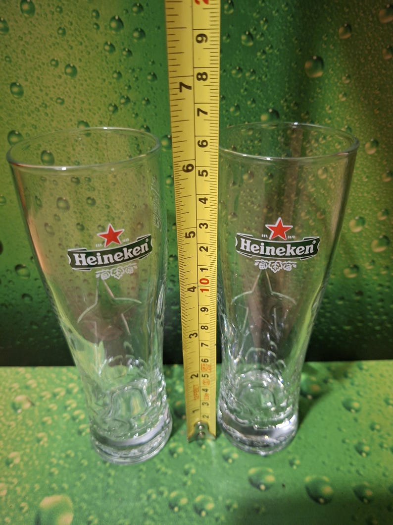 May include: Two clear Heineken beer glasses, each approximately 15 cm tall, with the Heineken logo and a star design. The glasses are on a green background with water droplets.