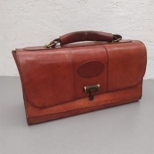 Puede incluir: Un maletín de cuero marrón vintage con asa y herrajes color latón. La bolsa rectangular presenta un cierre de solapa y un pequeño acento ovalado. El maletín está diseñado para transportar documentos y otros artículos.