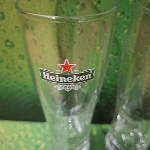 May include: Clear, tulip-shaped beer glass with the Heineken logo, featuring a red star and green lettering. The glass is empty and has a wide mouth, ideal for enjoying a cold beverage. The background is a green gradient.
