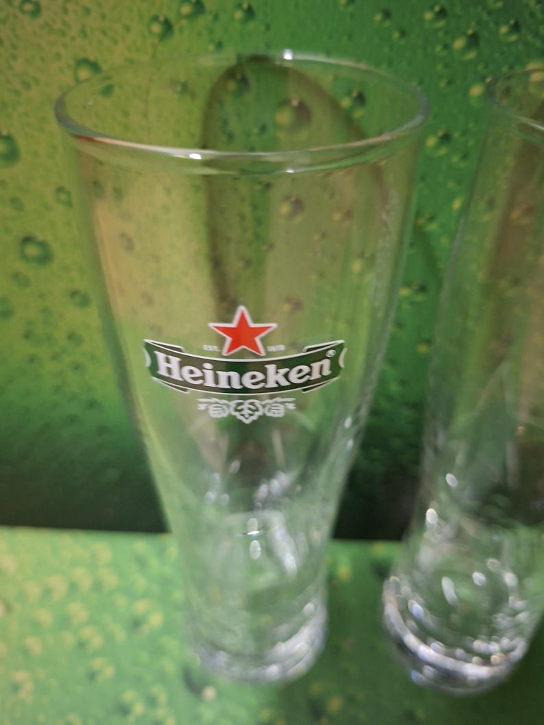 May include: Clear, tulip-shaped beer glass with the Heineken logo, featuring a red star and green lettering. The glass is empty and has a wide mouth, ideal for enjoying a cold beverage. The background is a green gradient.