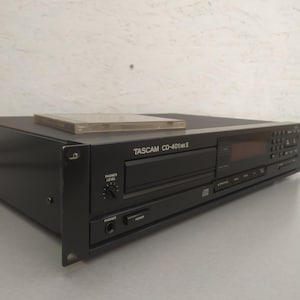 Könnte beinhalten: Schwarzer Tascam CD-401MKII CD-Player mit einer transparenten CD-Hülle oben. Der Player hat ein digitales Display, Knöpfe und einen Kopfhöreranschluss. Der Text "TASCAM CD-401MKII" ist auf der Vorderseite aufgedruckt.