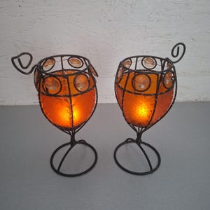 Peut inclure: Deux bougeoirs en forme de calice composés d'une structure en fil de fer brun foncé et de panneaux en verre orange. Chaque support présente une base circulaire, un pied et un bord orné de perles de verre rondes et d'une spirale en fil de fer.