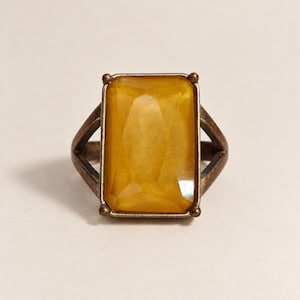 Può includere: Un anello con una grande pietra rettangolare sfaccettata color ambra incastonata in una montatura in metallo scuro e ossidato. La fascia presenta un design diviso che si collega ai lati dell'incastonatura, con una finitura metallica usurata.