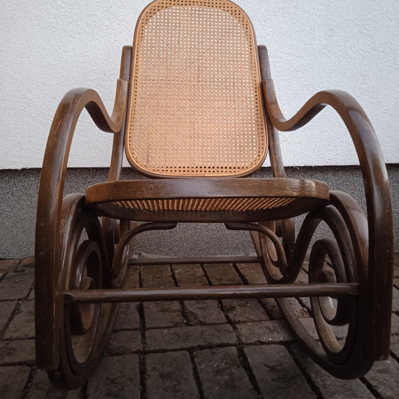 Peut inclure: Une chaise ber&ccedil;ante en bois brun avec un si&egrave;ge et un dossier en osier tress&eacute;. La chaise a un design incurv&eacute; avec des d&eacute;tails complexes.