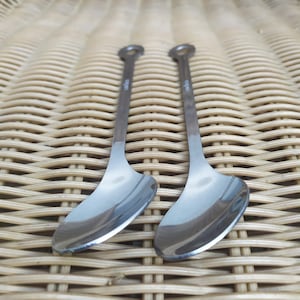 Könnte beinhalten: Zwei Servierlöffel aus Edelstahl mit einer abgerundeten Schüssel und einem langen, dünnen Stiel. Die Löffel haben eine glänzende, reflektierende Oberfläche und ein kleines kreisförmiges Detail am Ende des Stiels. Die Löffel dienen wahrscheinlich zum Servieren von Speisen.