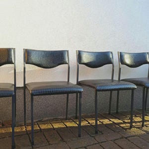 Peut inclure: Quatre chaises noires avec un siège en cuir gaufré crocodile et une structure en métal noir. Les chaises sont alignées en rangée contre un mur blanc.
