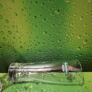 May include: A clear beer glass with the Heineken logo, including a red star and green details. The glass is positioned against a green backdrop with water droplets.