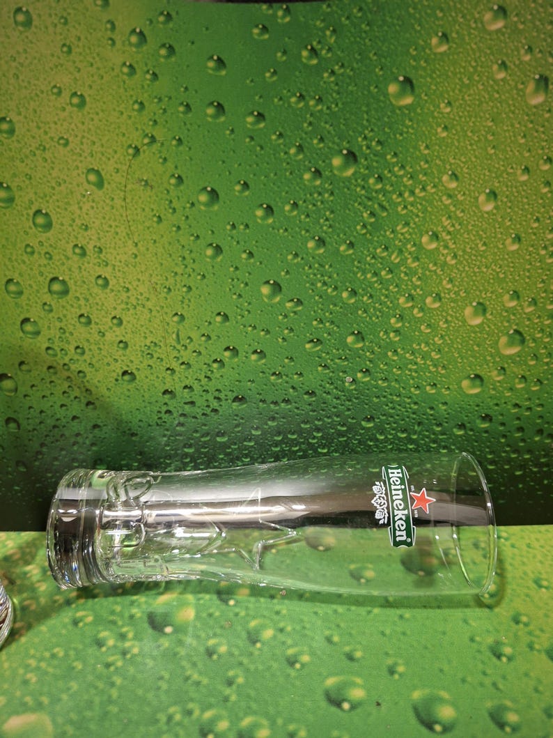 May include: A clear beer glass with the Heineken logo, including a red star and green details. The glass is positioned against a green backdrop with water droplets.
