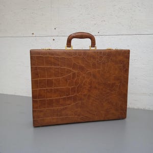 May include: A brown briefcase with a crocodile skin texture. The briefcase has a handle and gold-colored hardware. The case is rectangular and sits on a gray surface. The background is a white wooden wall.