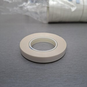 Könnte beinhalten: Eine Rolle cremefarbenes Abdeckband mit einem weißen Innenkern liegt auf einer grauen Oberfläche. Das Band ist kreisförmig, und eine Rolle ähnlichen Bandes ist im Hintergrund zu sehen, teilweise durch eine transparente Plastiktüte verdeckt.