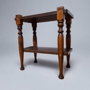 May include: A two-tiered wooden side table with a dark brown top and shelf. The table has four turned legs and is constructed with visible wood grain. The legs are a lighter brown colour than the top and shelf. The table is on a grey background.