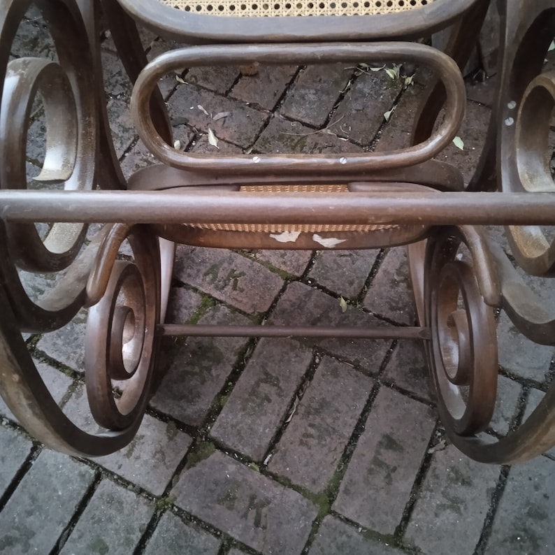 Peut inclure: Une chaise ber&ccedil;ante en bois brun avec un si&egrave;ge et un dossier en cannage. La chaise est pos&eacute;e sur un patio en briques.