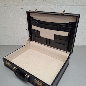 Peut inclure: Mallette en cuir noir ouverte, avec serrure à combinaison et garnitures dorées. L'intérieur est beige et comporte plusieurs poches. La mallette, avec une poignée, est conçue pour transporter des documents et des objets essentiels.