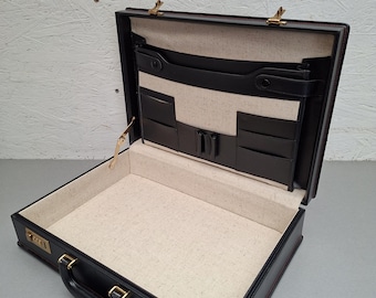 Op de afbeelding: Zwarte leren aktetas, opengeklapt met een beige binnenkant en meerdere vakken. De aktetas heeft een cijferslot en goudkleurige hardware. Ontworpen voor het dragen van documenten en heeft een handvat. Afmetingen zijn ongeveer 40 cm x 30 cm.