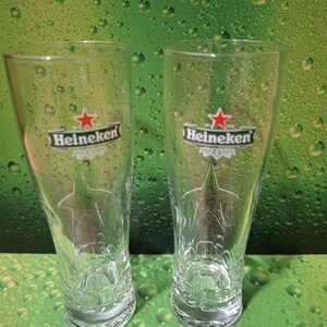 May include: Two clear Heineken beer glasses. Each glass features the Heineken logo with a red star and the brand name. The glasses have a star design etched into the glass. The background is green.