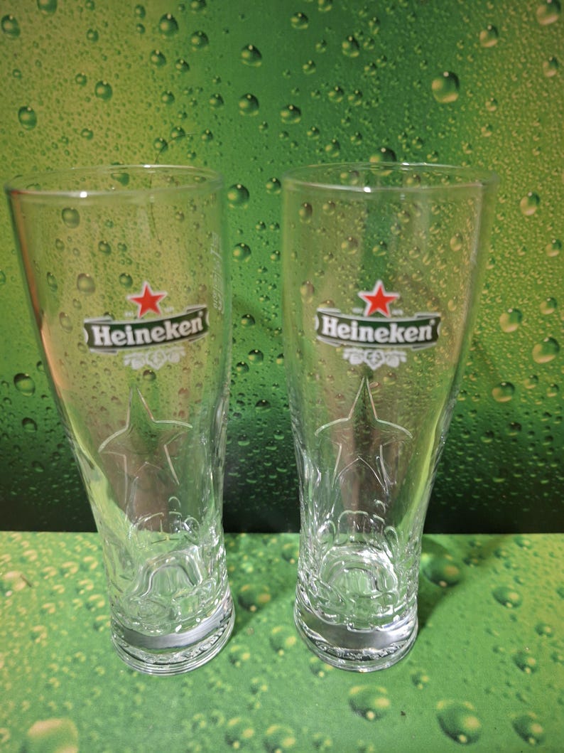 May include: Two clear Heineken beer glasses. Each glass features the Heineken logo with a red star and the brand name. The glasses have a star design etched into the glass. The background is green.