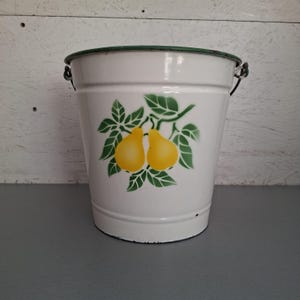 Pode incluir: Um balde de metal esmaltado branco com um gráfico pintado de duas peras amarelas rodeadas por folhas verdes na parte frontal. O balde tem uma borda verde-escura e uma alça de metal fixada nas laterais com pequenas argolas metálicas.