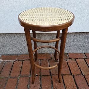 May include: A vintage wooden stool with a round woven cane seat. The stool has a dark brown finish and curved legs, with a circular footrest. The stool is approximately 28 inches tall.