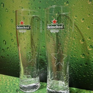 May include: Two clear, tall beer glasses featuring the Heineken logo and a star design. The glasses have a curved shape and are set against a green background with water droplets.
