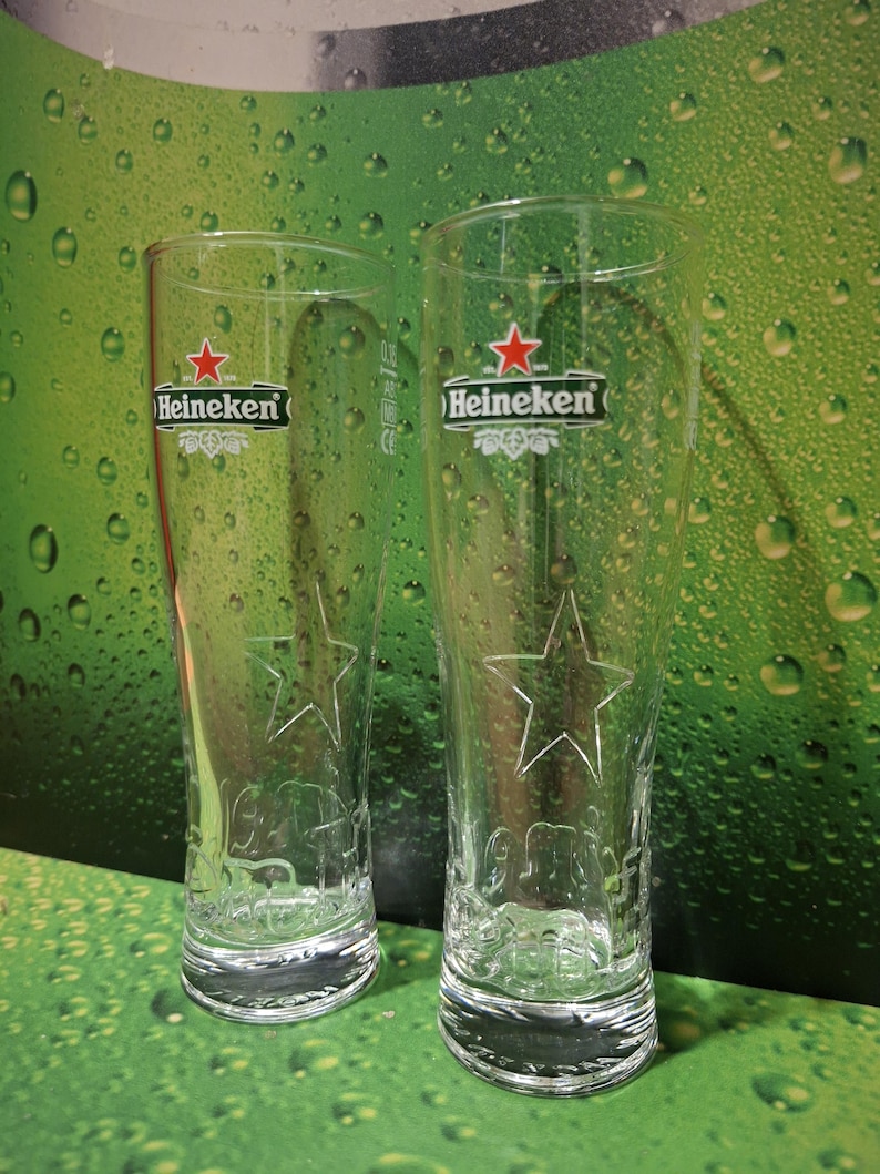 May include: Two clear, tall beer glasses featuring the Heineken logo and a star design. The glasses have a curved shape and are set against a green background with water droplets.