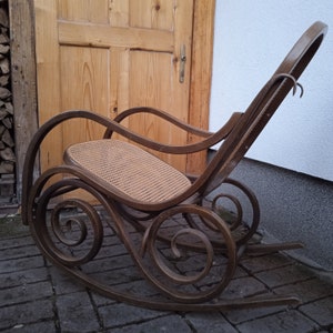 Peut inclure: Une chaise ber&ccedil;ante en bois avec un si&egrave;ge en cannage et un dossier incurv&eacute;. La chaise pr&eacute;sente des d&eacute;tails de volutes complexes sur les accoudoirs et les pieds.