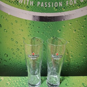 May include: Two clear Heineken beer glasses, each with the Heineken logo featuring a red star, are shown. They stand on a green surface with water droplets, against a backdrop with the words "Brewed with Passion for Quality."