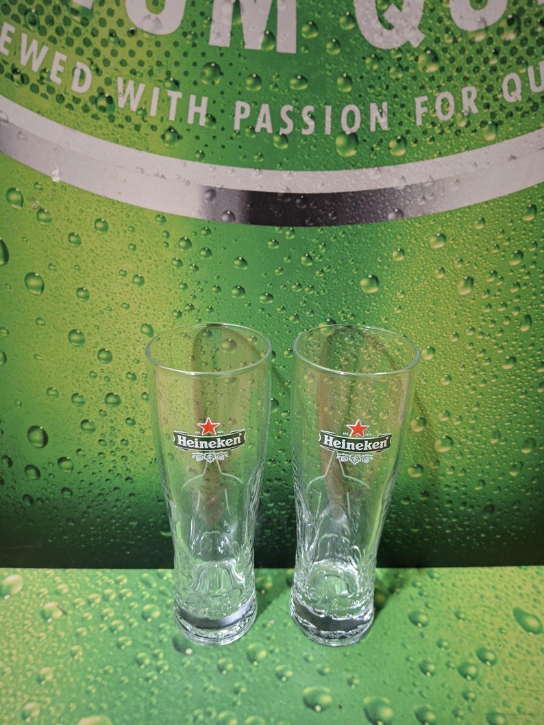 May include: Two clear Heineken beer glasses, each with the Heineken logo featuring a red star, are shown. They stand on a green surface with water droplets, against a backdrop with the words "Brewed with Passion for Quality."