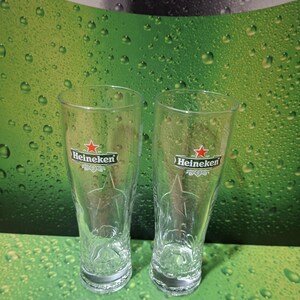 May include: Two clear, tulip-shaped beer glasses with the Heineken logo, featuring a red star and green text. The glasses have a textured design and sit against a green background with water droplets.