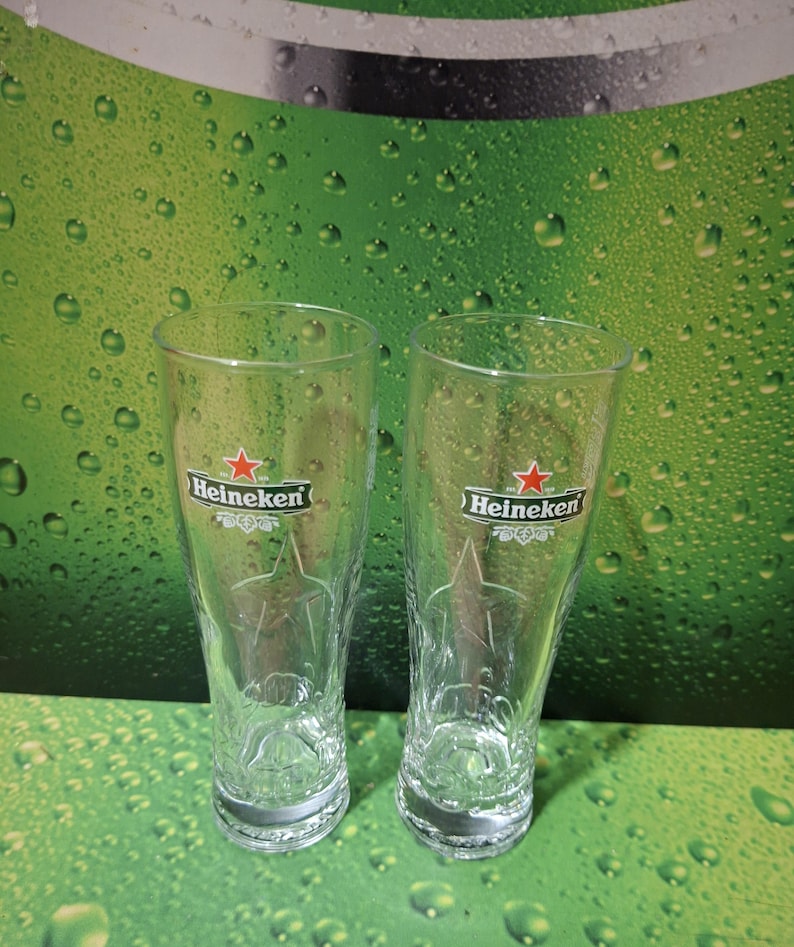May include: Two clear, tulip-shaped beer glasses with the Heineken logo, featuring a red star and green text. The glasses have a textured design and sit against a green background with water droplets.