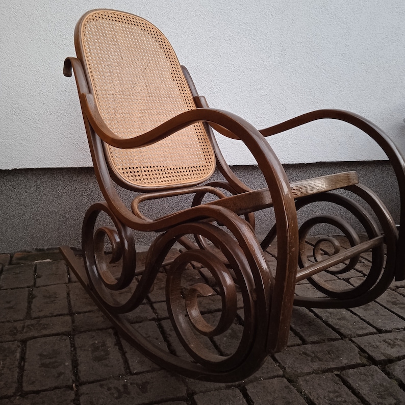 Peut inclure: Une chaise ber&ccedil;ante en bois brun avec un si&egrave;ge et un dossier en osier tress&eacute;. La chaise a un design incurv&eacute; avec des d&eacute;tails complexes et une base large.