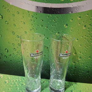 May include: Two clear Heineken beer glasses, each with the Heineken logo featuring a red star. The glasses are set against a green backdrop with water droplets and the words "WITH PASSION FOR".