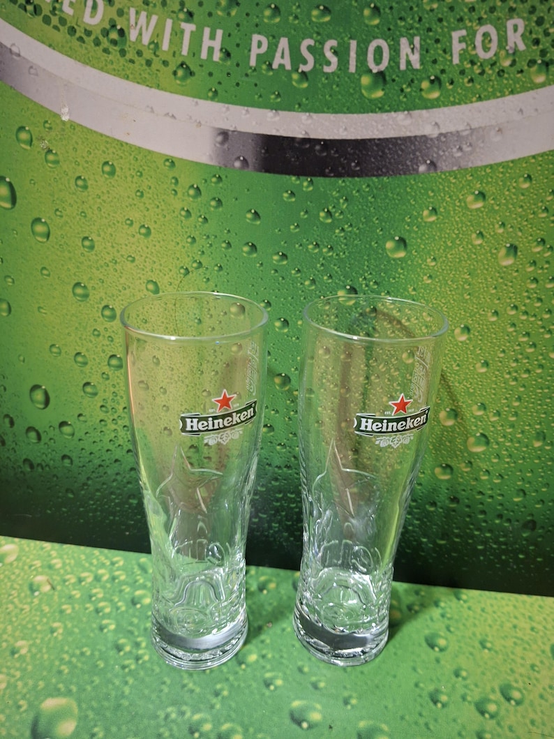 May include: Two clear Heineken beer glasses, each with the Heineken logo featuring a red star. The glasses are set against a green backdrop with water droplets and the words "WITH PASSION FOR".