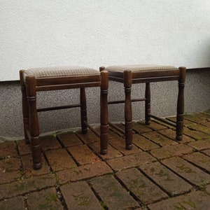 May include: Two wooden stools with a light brown fabric seat. The stools are sitting on a brick patio.