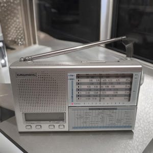 Puede incluir: Una radio portátil Grundig plateada con antena telescópica. La radio tiene una pantalla digital, un dial de sintonización y un selector de banda. La cara muestra información de frecuencia y zonas horarias mundiales. La radio tiene una correa de transporte.