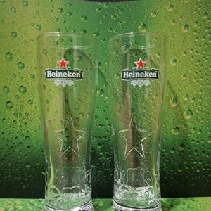 May include: Two clear, tall beer glasses featuring the Heineken logo and a star design. The glasses are empty and set against a green backdrop with water droplets.