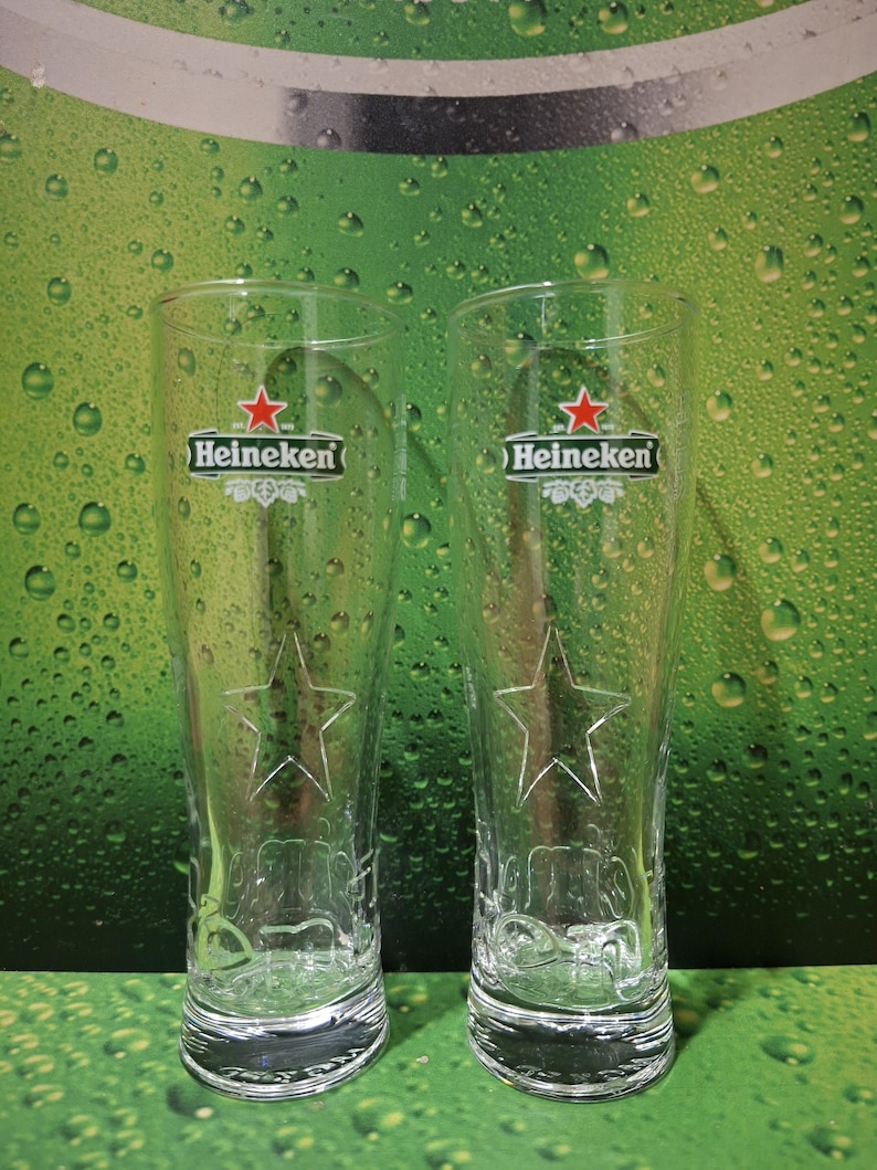 May include: Two clear, tall beer glasses featuring the Heineken logo and a star design. The glasses are empty and set against a green backdrop with water droplets.