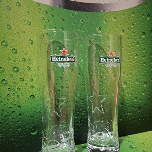 May include: Two clear, tall beer glasses featuring the Heineken logo and a star design. The glasses are empty, set against a green backdrop with water droplets and the text "PASSION FOR QUALITY".