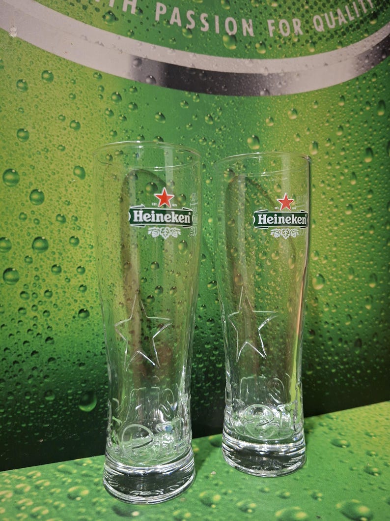 May include: Two clear, tall beer glasses featuring the Heineken logo and a star design. The glasses are empty, set against a green backdrop with water droplets and the text "PASSION FOR QUALITY".