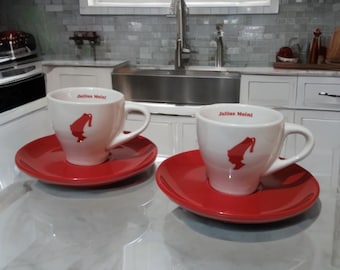 Peut inclure: Deux tasses à expresso blanches avec des soucoupes rouges. Chaque tasse porte l'inscription "Julius Meinl" et une silhouette rouge. Les tasses sont posées sur un comptoir.