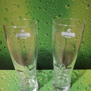 May include: Two clear Heineken beer glasses, empty, on a green background with water droplets. Each glass has the Heineken logo. The glasses are made of clear glass.