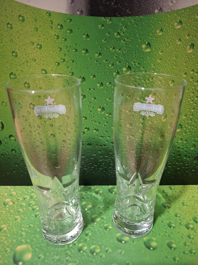May include: Two clear Heineken beer glasses, empty, on a green background with water droplets. Each glass has the Heineken logo. The glasses are made of clear glass.