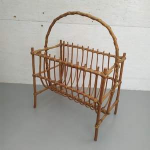 May include: A vintage, light brown rattan magazine rack with a curved handle. The rack features vertical slats and a woven base, supported by four legs. The design is simple and functional, suitable for storing reading materials.