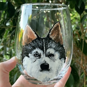May include: A clear glass wine glass with a hand-painted portrait of a black and white husky dog with brown ears. The dog's face is painted in detail, with a serious expression.